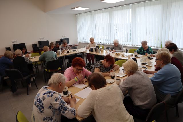 Na zdjęciu znajdują się kobiety siedzące przy złączonych w literę C stołach i rozwiązują zadania. Na stołach znajduje się również kawa, herbata i ciastka. W tle znajdują się okna i trzy komputery.