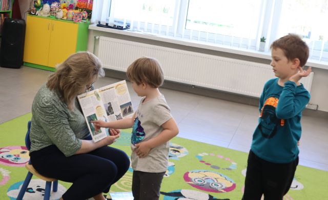 Na zdjęciu znajduje się siedząca kobieta z otwartą książką i pokazuje ilustracje dziewczynce, która stoi przed nią i wskazuje ilustracje palcem, kawałek za nią stoi chłopiec. W tle znajdują się okna.