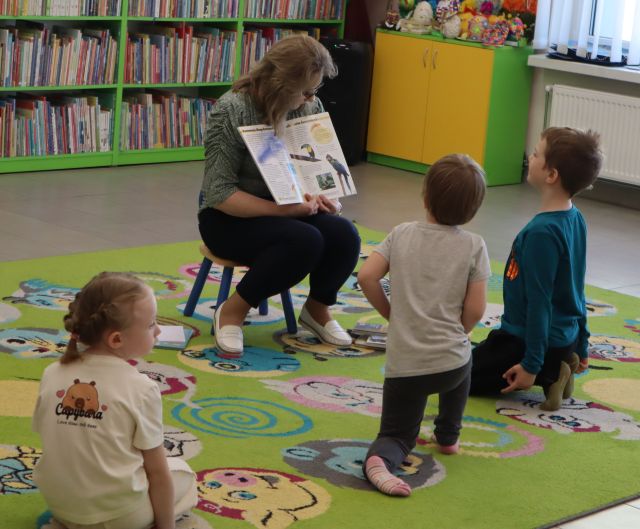 Na zdjęciu znajduje się kobieta na dywanie z otwartą książką i pokazuje ilustracje znajdującej się przed nią trójce dzieci. W tle znajdują się półki z książkami.