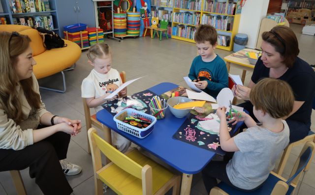Na zdjęciu znajduje się trojka dzieci i dwie kobiety przy stoliku, przy którym wycinają i kleją ptaki. W tle znajdują się półki z książkami oraz żółta, okrągła kanapa.