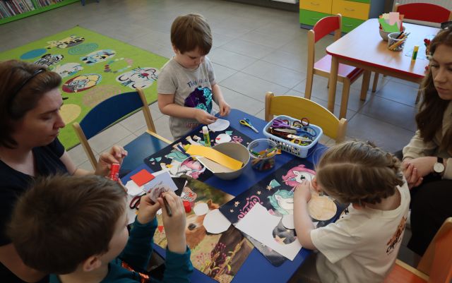 Na zdjęciu znajduje ise trójka dzieci przy stole wraz z dwójką kobiet, przy którym wycinają i kleją swoje prace. W tle znajduje się dywan, a po prawej stronie stolik.