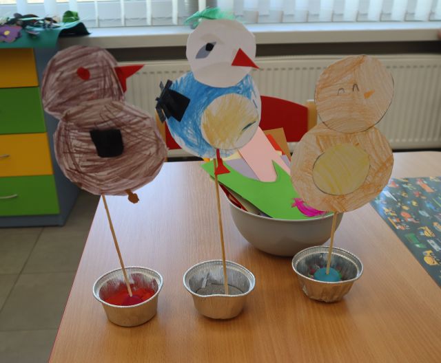 Na zdjęciu znajdują się papierowe ptaki na wykałaczce, stojące na stole, za nimi leży miseczka z kartkami. W tle znajdują się fragmenty okien i komoda.