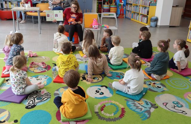 Na zdjęciu znajdują się dzieci na dywanie, a przed nimi siedzi kobieta z otwartą książką i pokazuje dzieciom ilustracje. W tle znajdują się półki z książkami, stolik.