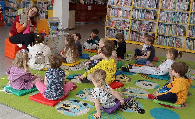 Na zdjęciu znajduje się piętnaście dzieci, które siedzą na dywanie i skierowane są w stronę siedzącej przed nimi kobietą, która pokazuje im ilustracje w książce. W tle znajdują się półki z książkami.