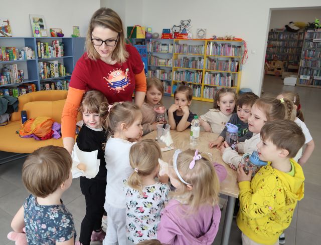 Na zdjęciu znajdują się dzieci oraz kobieta dookoła stołu . Kobieta pokazuje dziewczynce po lewej jajko, które leży na chusteczce. W tle znajdują się półki z książkami.