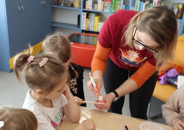 Na zdjęciu znajduje się kobieta, która nakłada pastę do zębów na szczoteczkę trzymaną przez dziewczynkę po lewej. Przy stoliku znajduje się trójka innych uczestników, a w tle znajdują się półki z grami, szafa i czerwony stolik multimedialny.