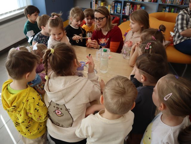 Na zdjęciu znajdują się dzieci dookoła stołu oraz kobieta, która trzyma jajko, które szczotkuje dziewczynka stojąca po lewej stronie. Zdjęcie zostało wykonane od góry i w tle znajdują się półki z grami.