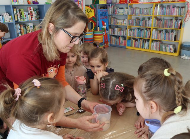 Na zdjęciu znajdują się dzieci oraz kobieta przy stole i obserwują jajko w plastikowym pojemniku. W tle znajdują się półki z książkami.