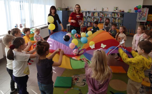 Na zdjęciu znajduje się piętnaście dzieci oraz dwie kobiety i wszyscy trzymają kolorową chustę, na której znajdują się balony. W tle znajdują się półki z grami, a po lewej stronie okna.