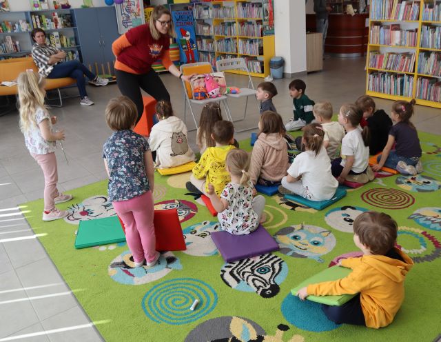 Na zdjęciu znajdują się dzieci na dywanie, oraz stojąca, pochylona kobieta przed nimi, na krzesełku obok niej siedzą dwie maskotki oraz książka. W tle znajdują się półki z książkami.