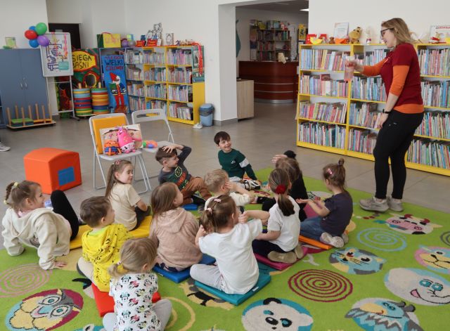 Na zdjęciu znajdują się dzieci siedzące na dywanie, po prawej stronie stoi kobieta i psika na nich wodą. W tle znajdują się półki z książkami.