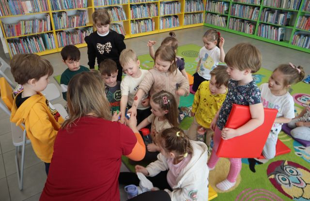 Na zdjęciu znajdują się dzieci stojące przed nachyloną po lewo kobietą, która rysuje uśmiech na ich ręce. Zdjęcie zostało wykonane od góry.