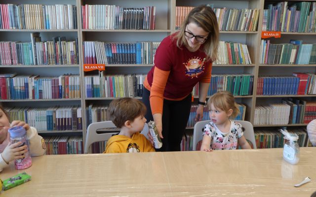 Na zdjęciu znajduje się kobieta, która daje do powąchania chłopcu, który siedzi przy stole pieprz. Przy złączonych stołach siedzą jeszcze dwie dziewczynki, stół jest owinięty folią. W tle znajdują się półki z książkami.