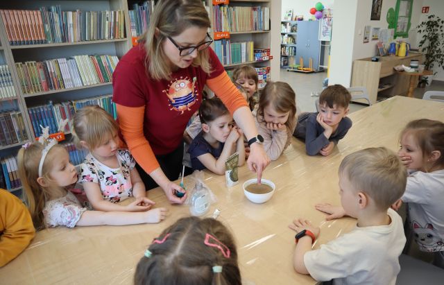 Na zdjęciu znajdują się dzieci stojące po obu stronach złączonych stołów, na środku stoi kobieta i wkłada palec do miski z wodą i pieprzem. W tle znajdują się półki z książkami.