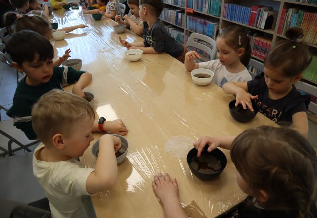 Na zdjęciu znajdują się dzieci siedzące przy złączonych stołach i wkładają palec do miski z wodą i pieprzem. W tle po prawo znajdują się półki z książkami.