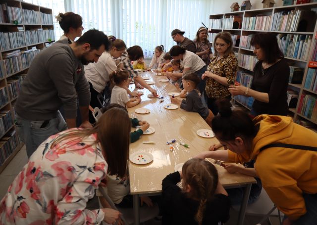 Na zdjęciu znajdują się dzieci wraz z rodzicami przy złączonych stołach i nakładaj farbę na talerzyki leżące przed nimi. W tle znajdują się okna, a po prawej i lewej stronie półki z książkami.