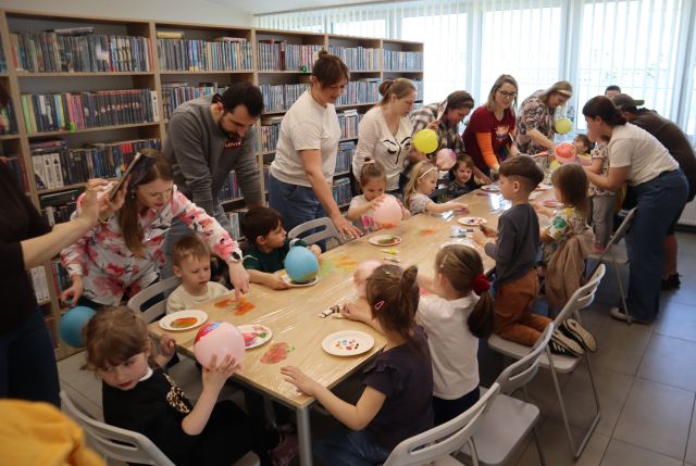 Na zdjęciu znajdują się dzieci wraz z rodzicami stojącymi za nimi przy złączonych stołach i za pomocą balonów odbijają farbę na folii na stole. W tle znajdują się półki z książkami i okna po prawej stronie.