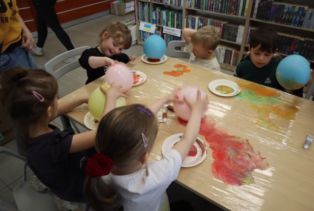 Na zdjęciu znajduje się piątka dzieci przy złączonych stołach i za pomocą balona odbijają farbę na folii rozłożonej na stole. Zdjęcie zostało wykonane od góry, a w tle znajdują się półki z książkami.