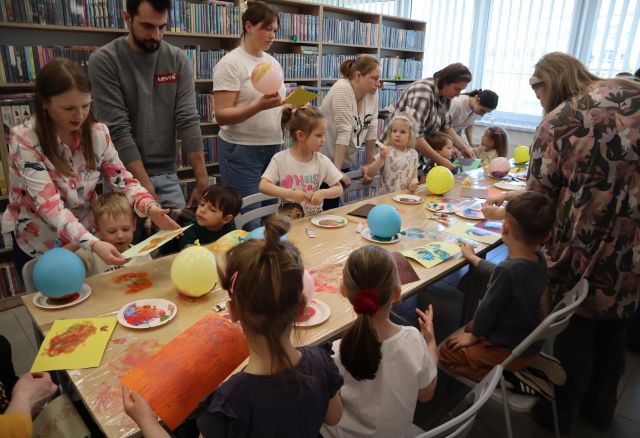 Na zdjęciu znajdują się dzieci odbijające farbę na folii znajdującej się na złączonych stołach, za dziećmi stoją rodzice i pomagają odbić wzór z folii na kolorowej kartce. W tle znajdują się półki z książkami, a po prawej stronie okna.