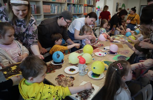 Na zdjęciu znajdują się dzieci wraz z rodzicami przy złączonych stołach i odbijają na folii za pomocą balonów farbę. W tle znajdują się półki z książkami.