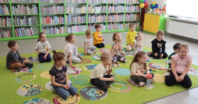 Na zdjęciu znajduje się czternaście dzieci siedzących na dywanie. W tle znajdują się półki z książkami.