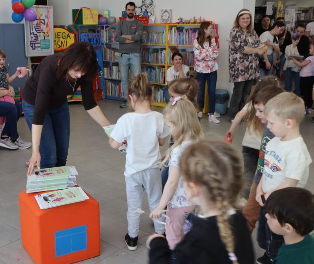 Na zdjęciu znajdują się dzieci stojące w kolejce dopochylonej kobiety, która wręcza każdemu książkę. W tle znajdują się półki z książkami oraz rodzice.