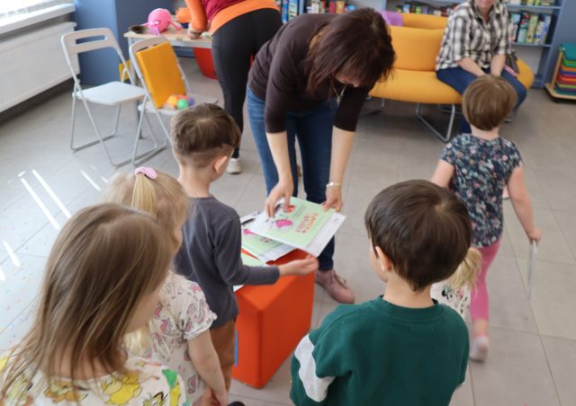 Na zdjęciu znajduje się piątka dzieci stojące przy kobiecie, która daje każdemu po książce. W tle znajdują się dwa krzesełka, stolik.