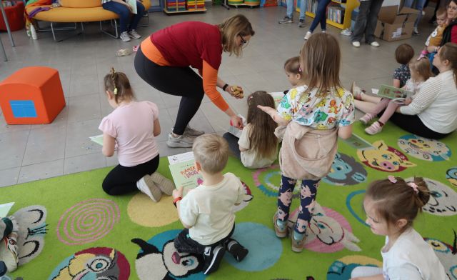 Na zdjęciu znajdują się dzieci na dywanie oraz kobieta, która się nachyla i przykleja każdemu naklejkę. W tle znajduje się pufa.