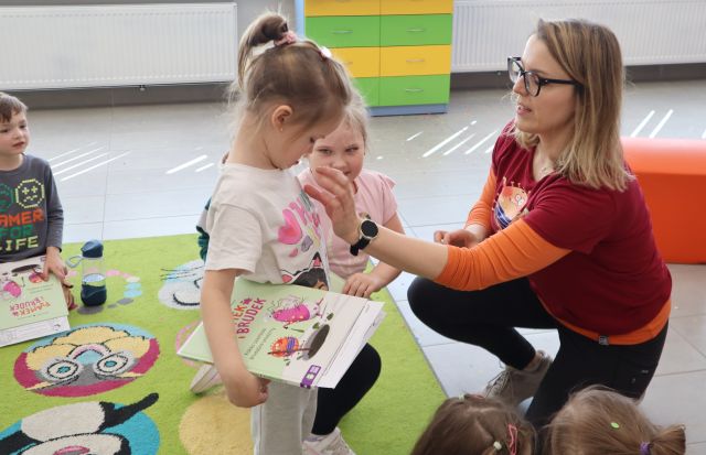 Na zdjęciu znajduje się kobieta naklejająca dziewczynce na bluzkę naklejkę, dziewczynka trzyma w ręce książkę. Koło nich znajduje się czwórka dzieci, a w tle znajduje się szafka.