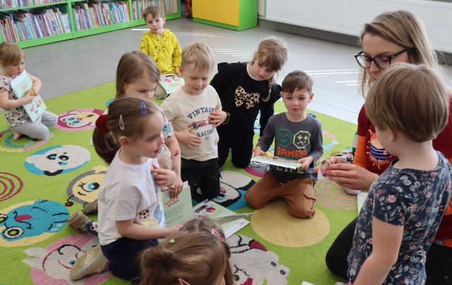 Na zdjęciu znajdują się dzieci na dywanie, a po prawej stronie kobieta przyklejająca dziewczynce na bluzkę naklejkę. W tle znajdują się półki z książkami i szafka.