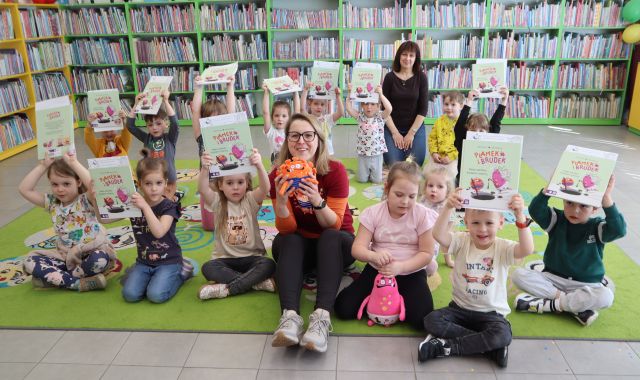 Na zdjęciu znajduje się piętnaście dzieci na dywanie, które trzymają podniesione do góry książki, wraz z nimi do zdjęcia siedzą dwie kobiety, jedna z nich, siedząca na przodzie ma w ręce maskotkę. W tle znajdują się półki z książkami.