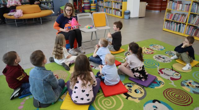 Na zdjęciu znajduje się dziewięcioro dzieci, które siedzą na dywanie, a przed nimi siedzi kobieta i trzyma w ręce dwie maskotki. W tle znajdują się półki z książkami.