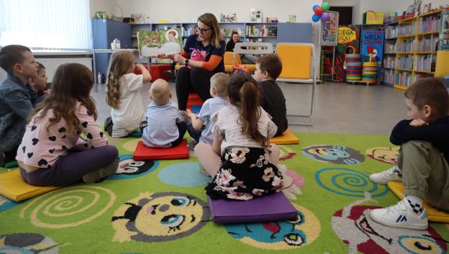 Na zdjęciu znajdują się dzieci siedzące na dywanie, a przed nimi siedzi kobieta z otwartą książką. W tle znajdują się półki z książkami.
