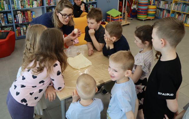Na zdjęciu znajdują się dzieci dookoła stolika wraz z kobietą, która pokazuje im jajko. W tle znajdują się półki z książkami.