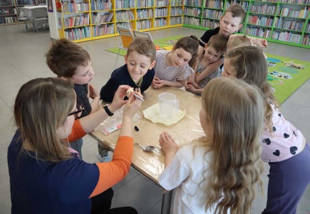 dzieci stoją wokół stołu, obok kuca kobieta trzymająca w dłoniach jajko