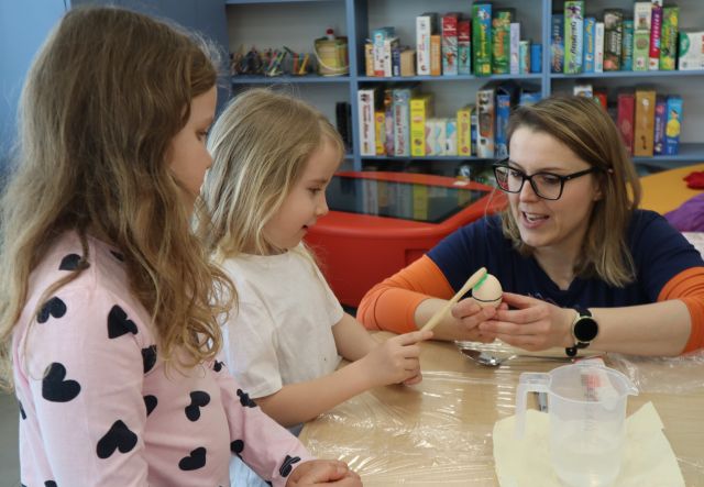 dziewczynka czyściu jajko szczoteczką, pozostałe dzieci stoją wokół stołu i się przypatrują, obok nich kuca kobieta