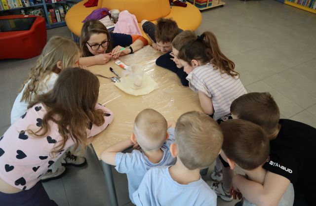 Na zdjęciu znajdują się dzieci oraz kobieta, oparci o stół i przyglądają się jajku, które leży w plastikowym pojemniczku na środku stołu. Zdjęcie zostało wykonane od góry i w tle znajduje się okrągłe siedzisko oraz stolik multimedialny.