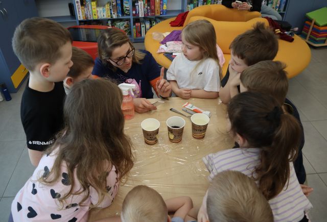Na zdjęciu znajdują się dzieci wraz z kobietą dookoła stołu, na którym leżą trzy kubeczki. W tle znajdują się półki z książkami i żółte okrągłe siedzisko.