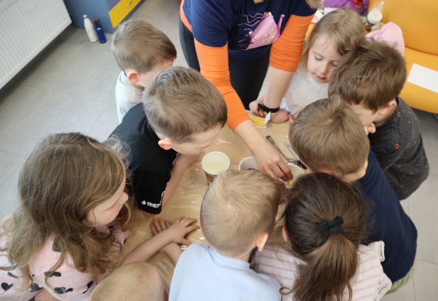 Na zdjęciu znajdują się dzieci przy stoliku oraz kobieta, która wkłada do jednego kubeczka papierek lakmusowy. Zdjęcie zostało wykonane od góry.