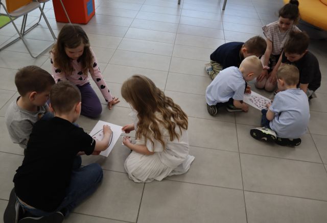 Na zdjęciu znajdują się dzieci siedzące na podłodze w dwóch grupach i mają przed sobą kartki z narysowanym zębem.