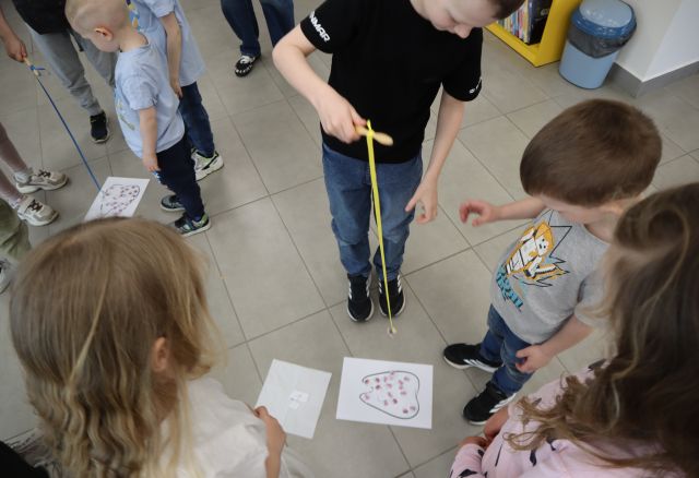 Na zdjęciu znajduje się grupka dzieci stojąca dookoła kartki z narysowanym zębem i za pomocą magnesu na sznurku łapią z kartki bakterie. Zdjęcie zostało wykonane od góry.