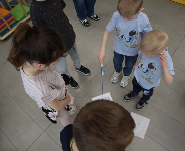Na zdjęciu znajdują się dzieci stojące dookoła kartki i łapią bakterie za pomocą magnesu na sznurku. Zdjęcie zostało wykonane od góry.