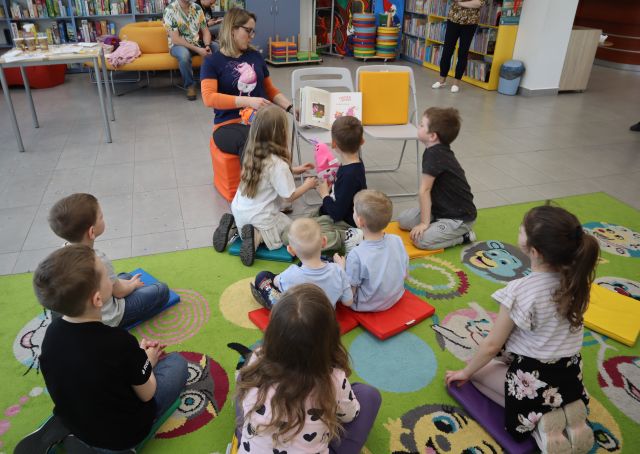 Na zdjęciu znajdują się dzieci na dywanie, przed nimi siedzi kobieta i obok niej na krześle stoi otwarta książka. W tle znajdują się półki z książkami, stolik.