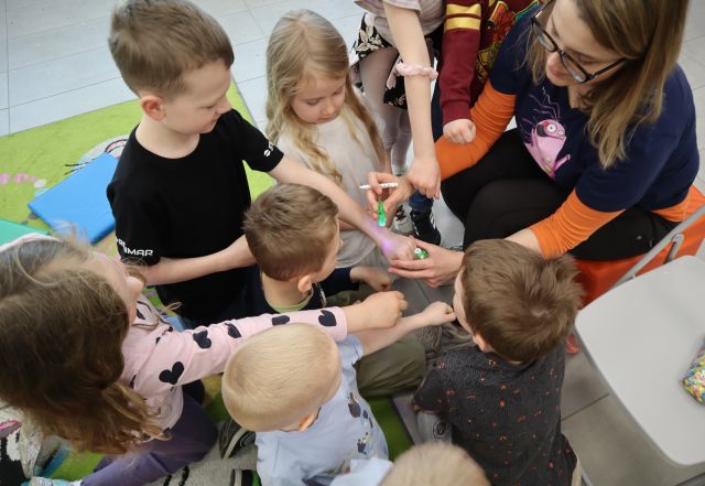 Na zdęciu znajdują się dzieci stojące przy kobiecie z wyciągniętymi rękami w stronę kobiety, która rysuje na ich ręce uśmiechy, które widać pod światłem UV. Zdjęcie zostało wykonane od góry.