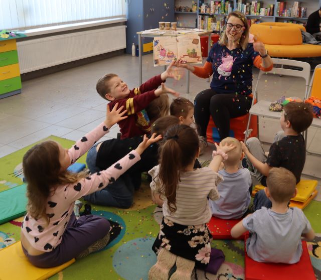 Na zdjęciu znajdują się dzieci na dywanie z wyciągniętymi rękami, przed nimi siedzi kobieta, która w jednej ręce trzyma otwartą książkę, a drugą ręką sypie na dzieci konfetti. W tle znajdują się okna, szafki.