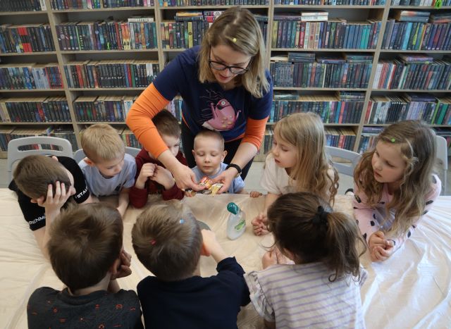 Na zdjęciu znajdują się dzieci stojące po obu stronach złączonych stołów, które są owinięte folią. Pomiędzy nimi stoi kobieta i trzyma w ręce opakowanie pieprzu, a na stole leży mydło. W tle znajdują się półki z książkami.