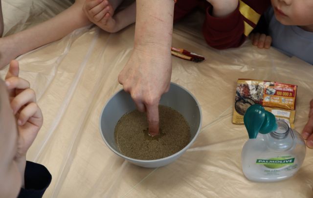Na zdjęciu znajduje się miseczka z wodą i pieprze, do której kobieta wkłada palec. Po prawej stronie leży mydło w płynie oraz opakowanie pieprzu. Dookoła kobiety znajdują się dzieci. Zdjęcie zostało wykonane od góry.