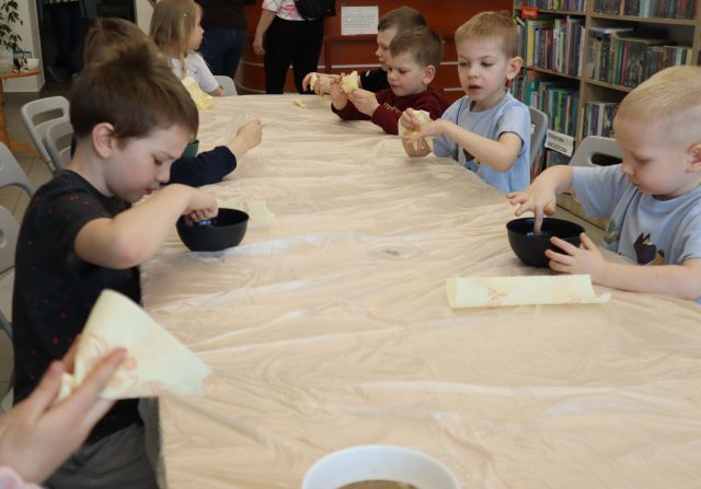 Na zdjęciu znajdują się dzieci, które siedzą przy złączonych stołach owiniętych folią, przed sobą mają miseczki z wodą, do których wkładają palec.