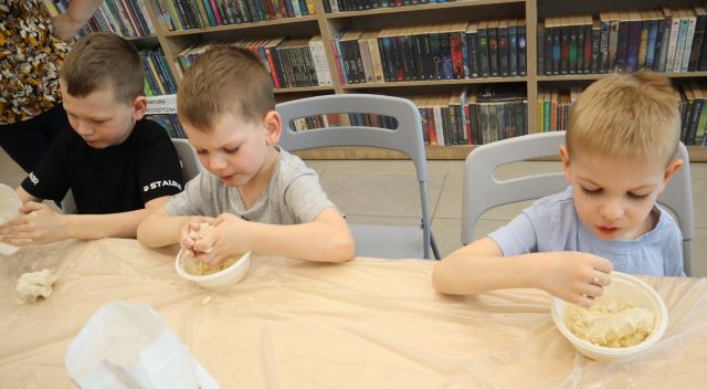 Na zdjęciu znajduje się trójka dzieci, która przy złączonych stolikach, owiniętych folią robią w miseczkach ciastolinę. W tle znajdują się półki z książkami.