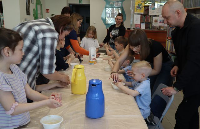 Na zdjęciu znajdują się dzieci przy złączonych stołach oraz piatka dorosłych, która pomaga dzieciom w zrobieniu ciastoliny, na stoliku stoja dwa termosy oraz mąka.